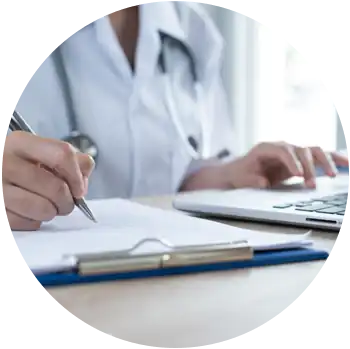 A light-skinned doctor is filling out paperwork on a clipboard with a pen while using a laptop.