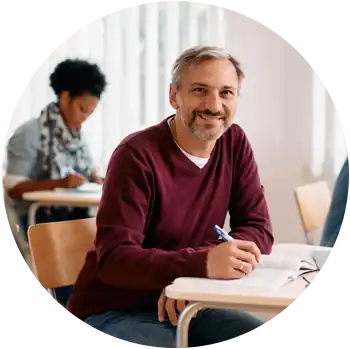 A medium skin toned man with salt and pepper hair sits at a desk in a classroom while holding a pen.