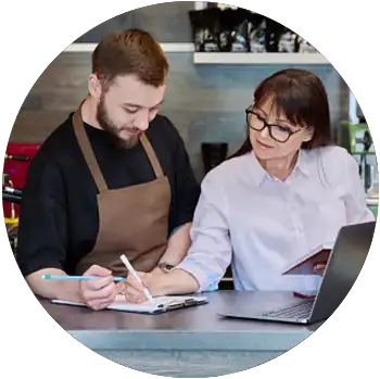 A white man and woman stand behind a counter at a cafe and look at a clipboard with paperwork. 